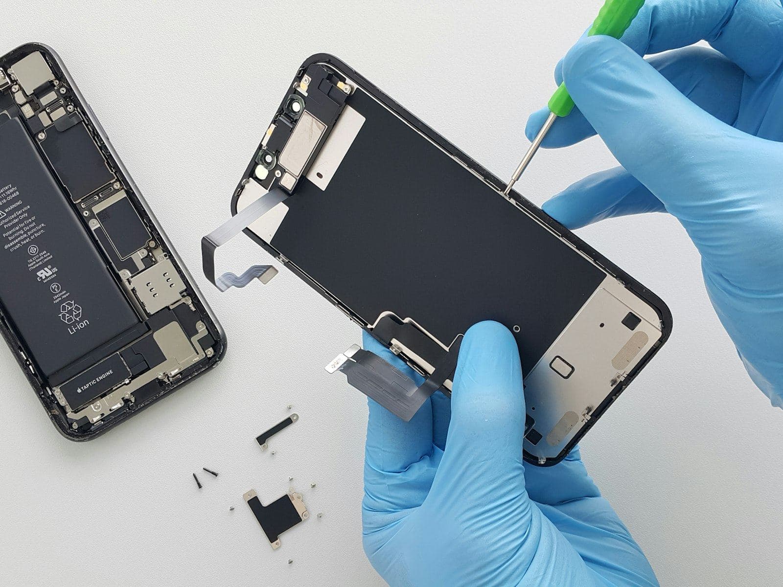 Mãos de luva azul abrindo um celular sobre a bancada, com a bateria e a placa à mostra